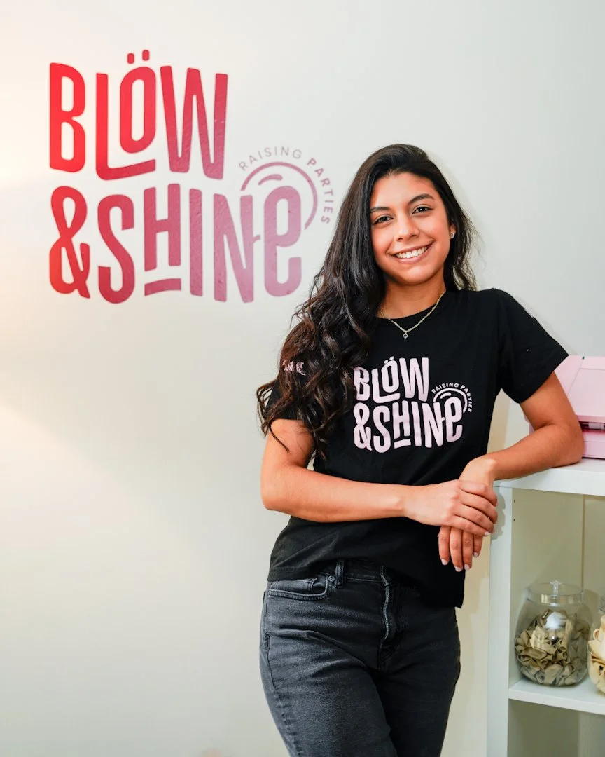 A young woman stands smiling next to a white wall with a logo that says 'BLOW & SHINE' and 'RAISING PARTIES'. She wears a black t-shirt with the same logo and has long, dark, wavy hair. There are pink boxes on a white shelf beside her.
