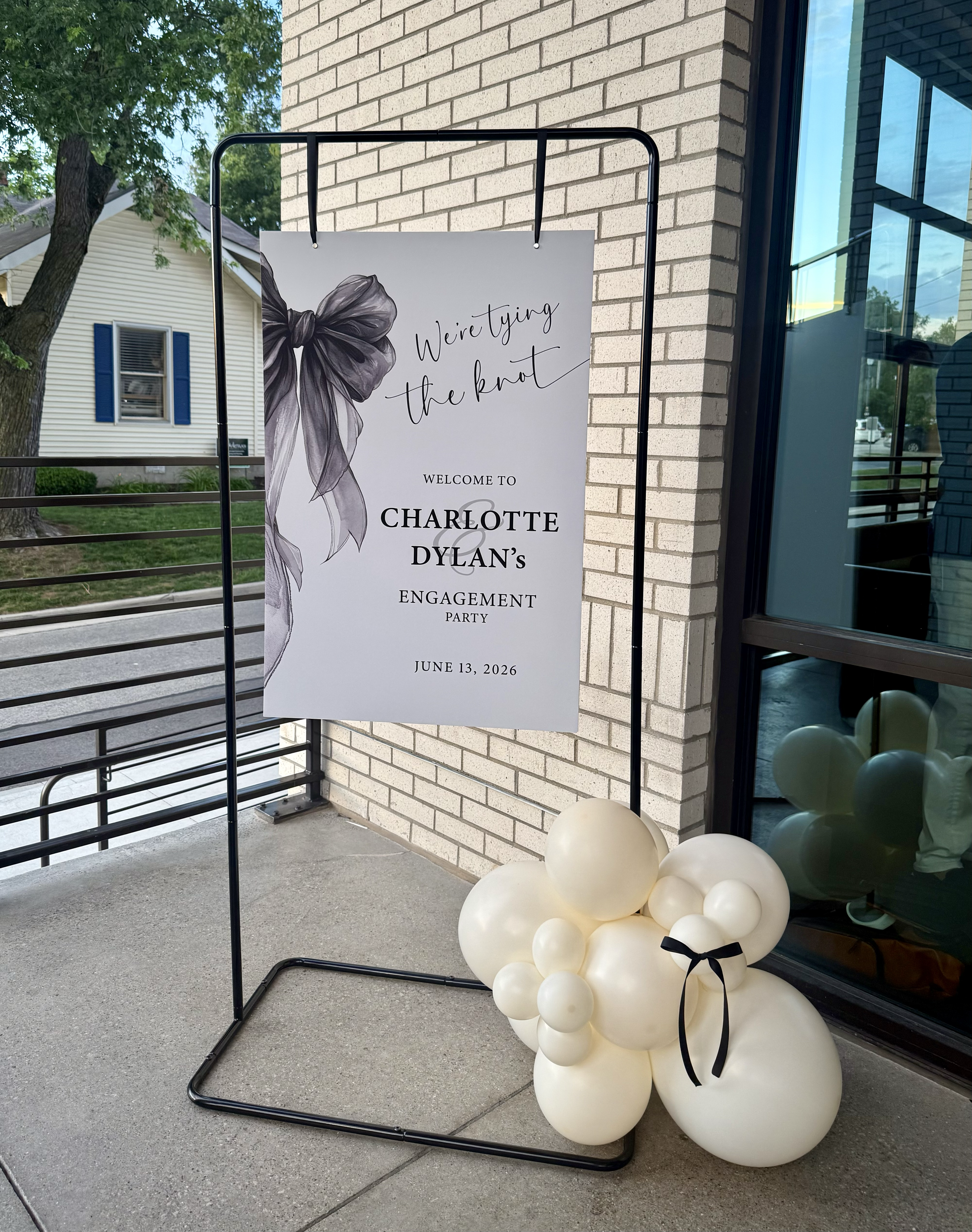 A decorative sign for engagement party with a black and white bow illustration and elegant script text, placed outdoors on a black metal stand near a building with brick wall and large glass window, with balloon decorations at the base.