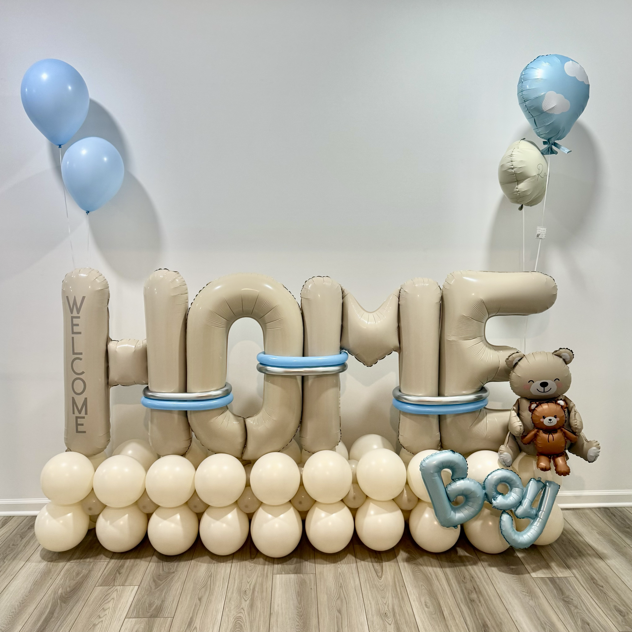 Balloon display spelling out the word 'WELCOME' with balloons in various shades of blue and beige, some with cloud and bear designs, on a wooden floor and plain wall background.