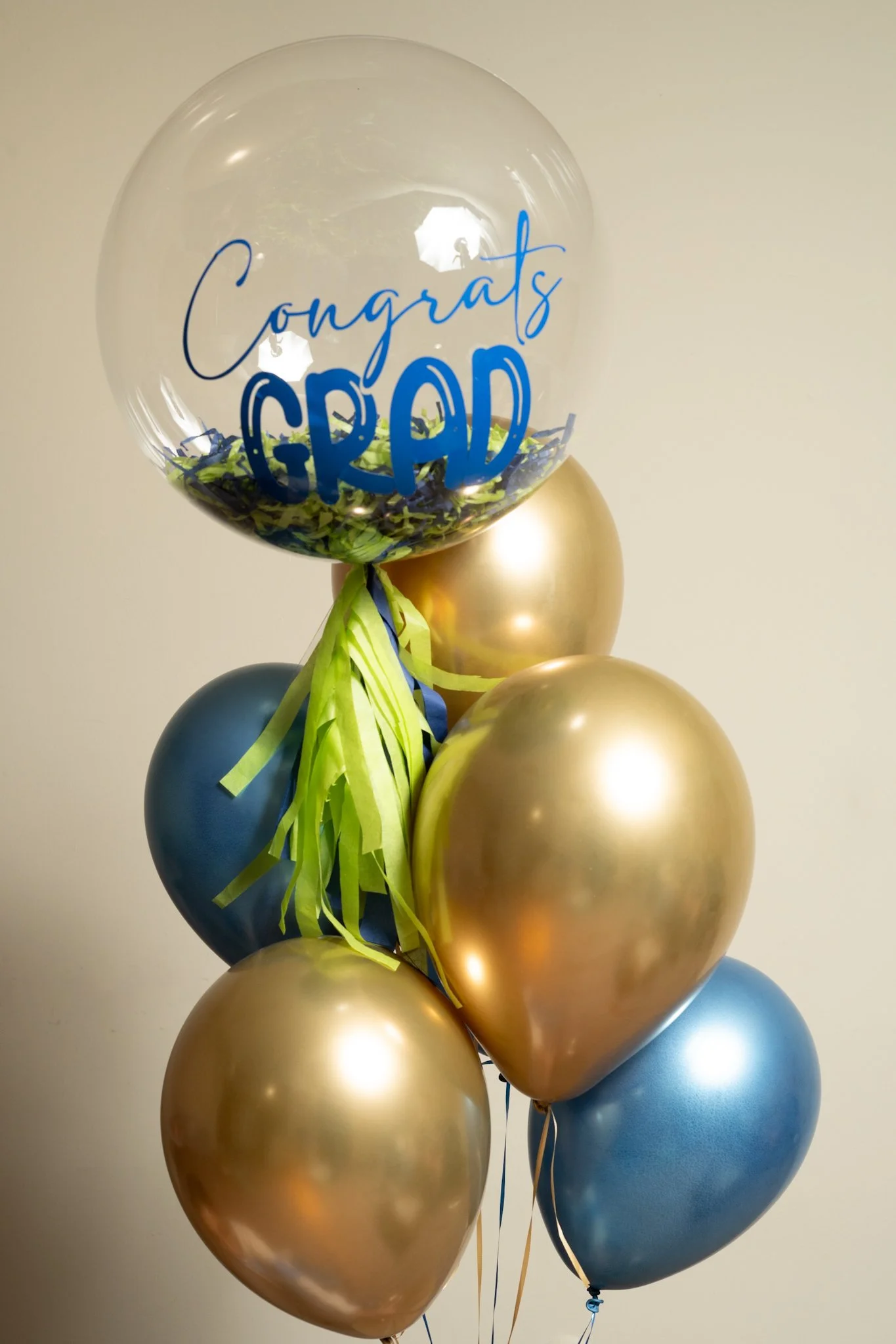 A bouquet of balloons with a large transparent balloon that has 'Congrats Grad' written on it, surrounded by gold and blue metallic balloons and green and blue ribbon streamers.