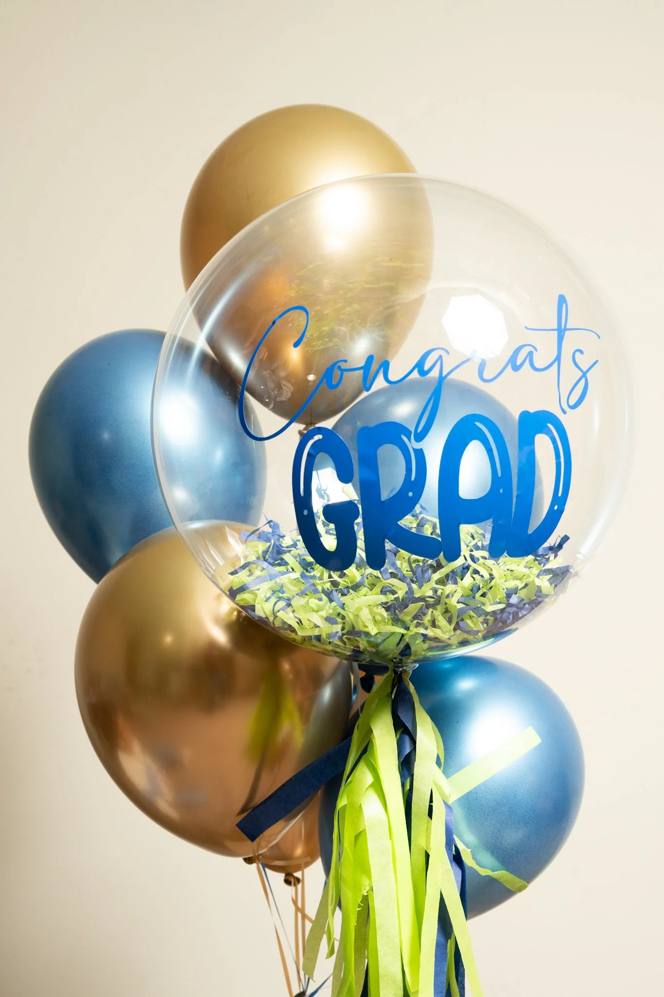 A bunch of metallic gold and blue balloons with a transparent balloon that says 'Congrats Grad' in blue text, tied together with green and blue tassels.