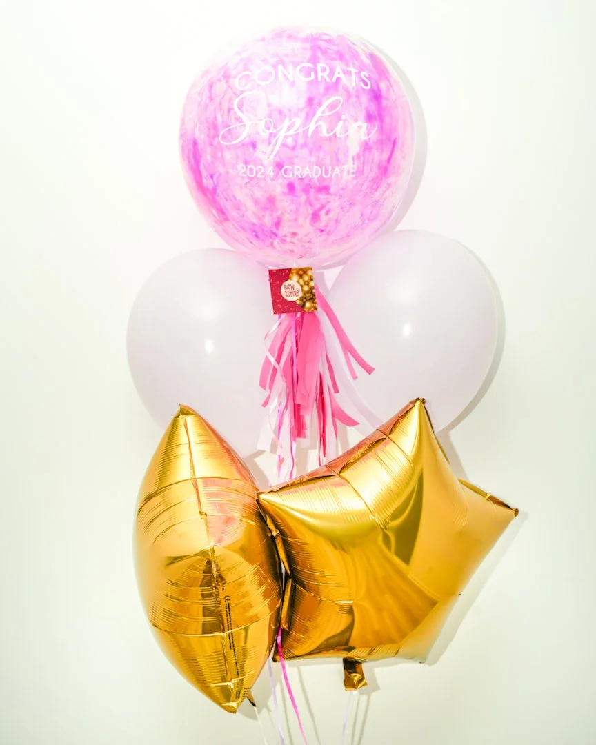 A celebration balloon arrangement with a large pink marbled balloon that has white text saying "Congrats Sophie 2024 Graduate," two white balloons, and three gold star-shaped balloons.