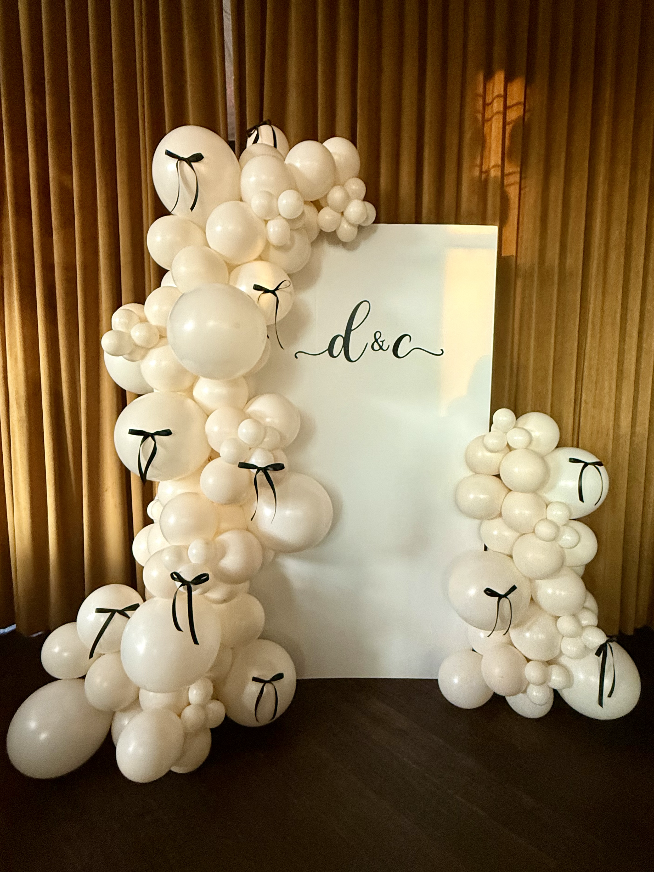A engagement balloon backdrop with white balloons and black ribbon accents surrounding a white board with 'd & c' written in black script.