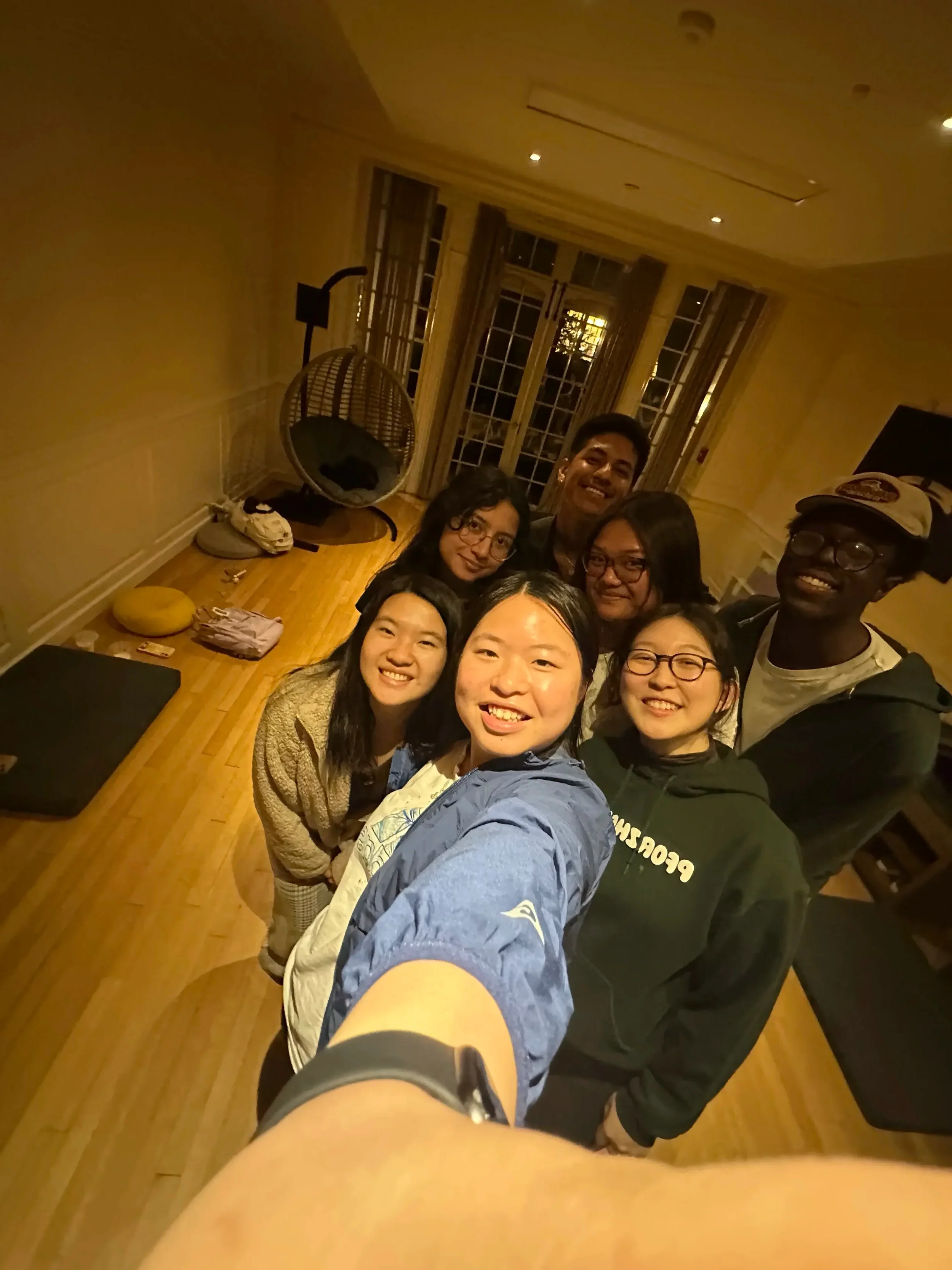 Group of seven diverse young adults taking a selfie in a warmly lit room with wooden floors and large windows.