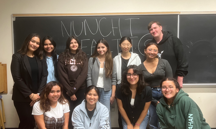 Group of eleven students and a teacher in a classroom standing in front of a blackboard with words 'NUNCHI' and 'MENTO' written on it.