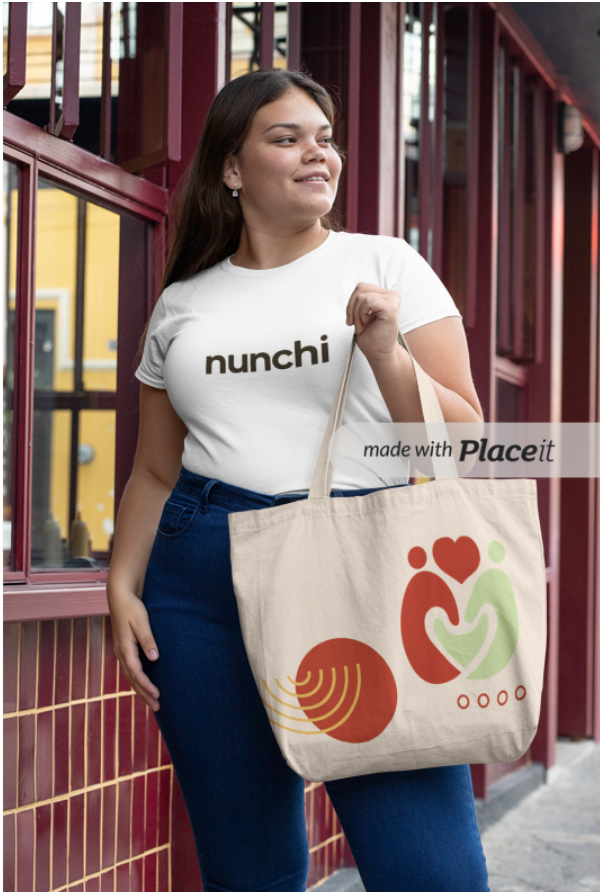 A young woman with long brown hair standing outdoors, holding a large beige tote bag with colorful abstract designs, wearing a white t-shirt with the word 'nunchi', and dark blue high-waisted jeans, smiling and looking off to the side near a maroon metal railing.