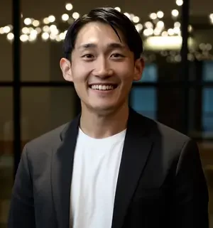 A smiling man in a blazer and white shirt standing indoors at night with city lights in the background.