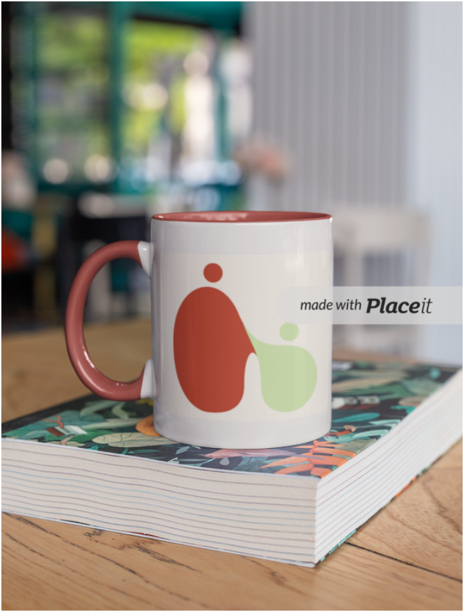 A white mug with a red interior and handle, featuring abstract green and red figures, sits on a large, colorful, patterned book on a wooden table. The background is blurred with a bright indoor space visible.