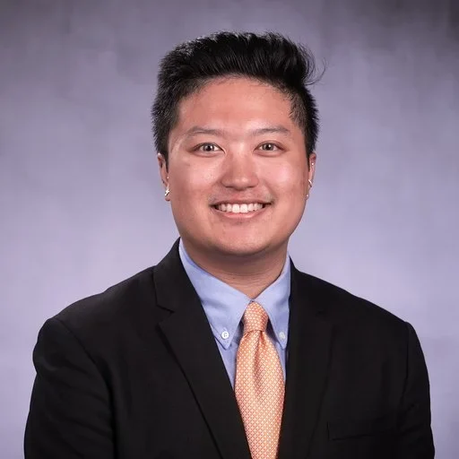 Professional portrait of a young man in a suit and tie, smiling, with a plain gradient background.