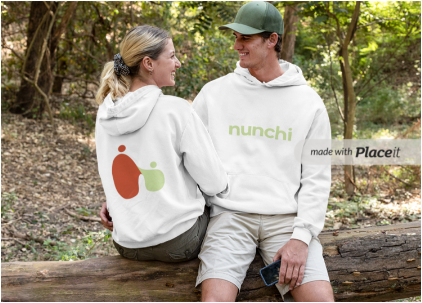 A man and woman sit on a log in a wooded area, smiling at each other. They wear white hoodies with minimalist designs and text. The woman’s hoodie has a colorful abstract logo on the back, and the man’s hoodie says "nunchi." He holds a smartphone in his right hand.