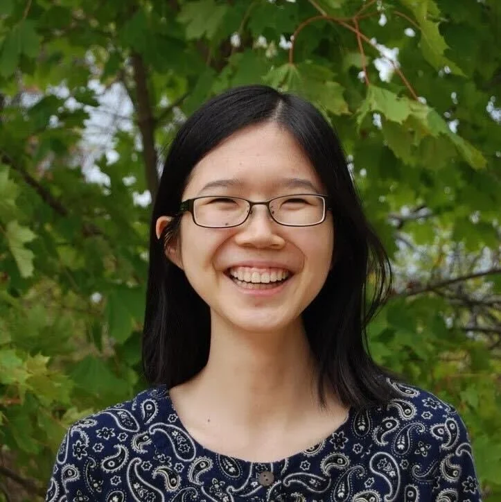 A man with glasses in a dark blue suit and white shirt, smiling outdoors with greenery in the background.