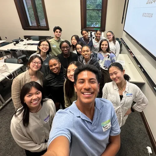 A group of young adults smiling for a selfie in a classroom with tables, computers, and large windows. There are about twelve people, with a man in the front taking the selfie.