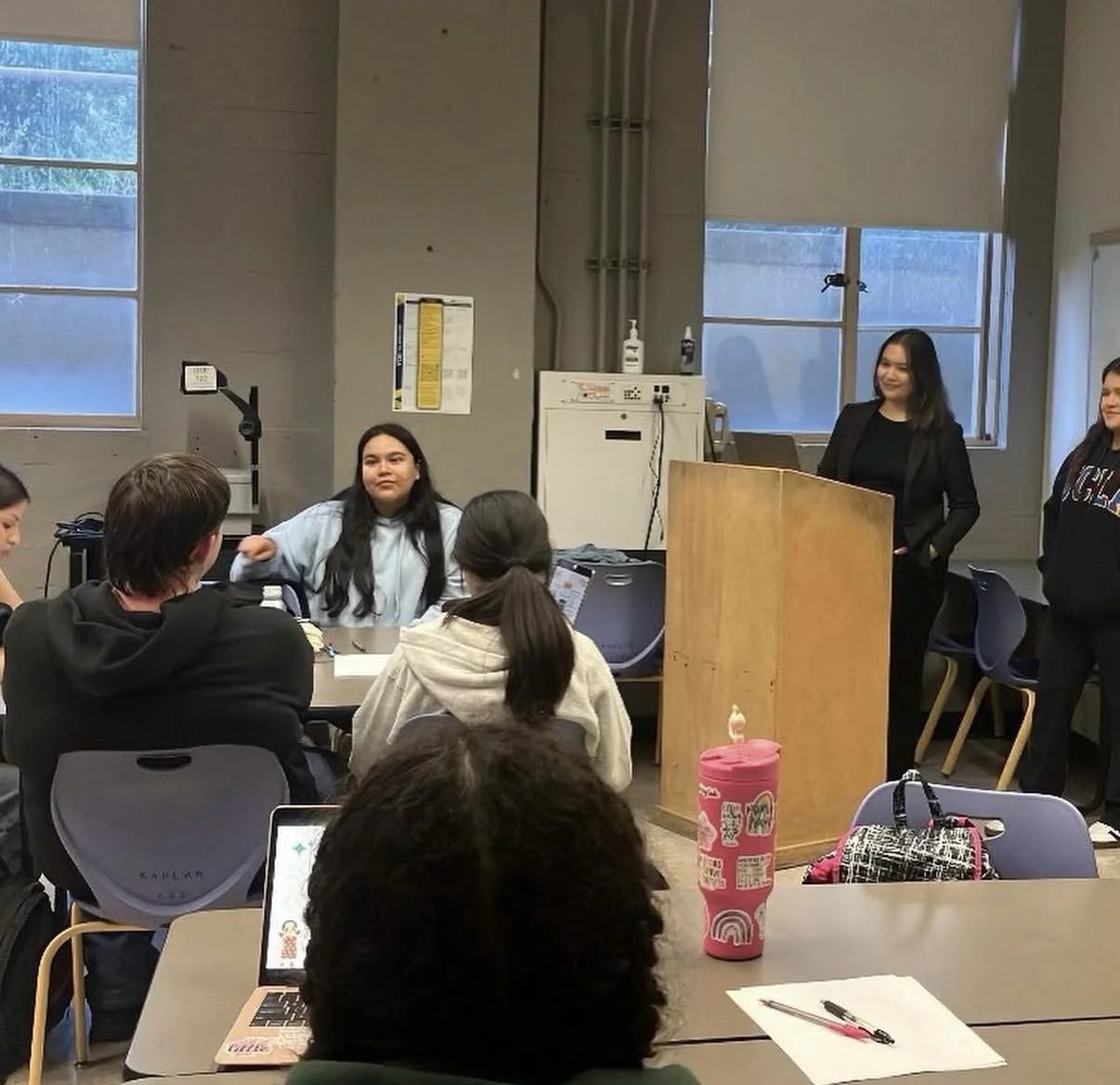 A classroom or meeting room with students and two women standing at the front, one behind a wooden podium, the other beside her. Students are sitting at tables, some with laptops and notebooks. The room has large windows, and a pink water bottle and a backpack are on the table.