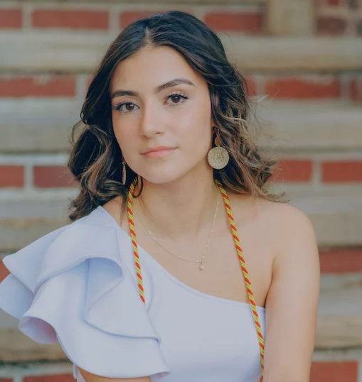 A young woman with wavy brown hair and light makeup, wearing a white top with a ruffled shoulder, a gold necklace, and large circular earrings, standing outdoors against a brick wall.