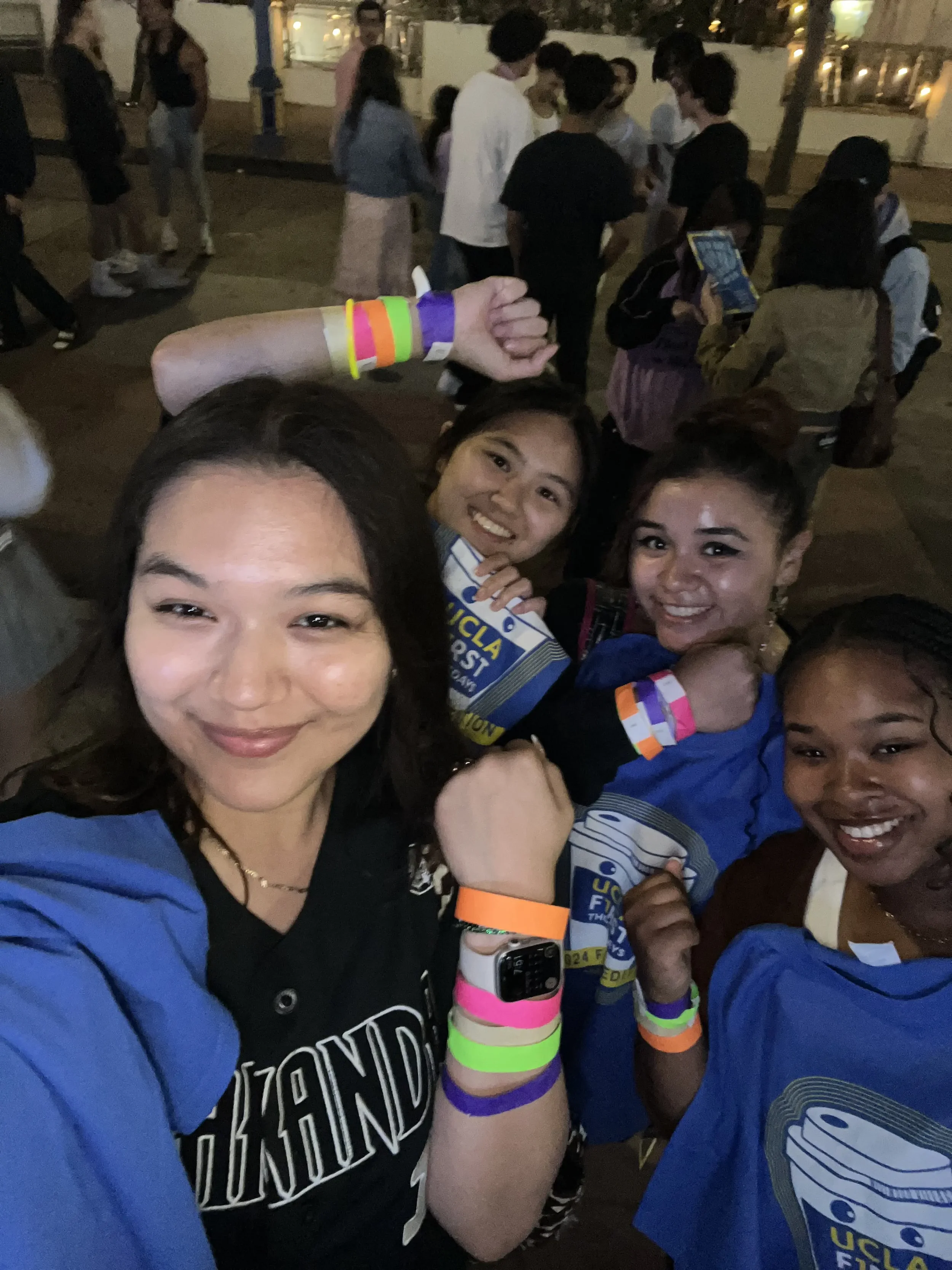 A group of smiling women taking a selfie at a nighttime outdoor event, some wearing colorful wristbands and event t-shirts.