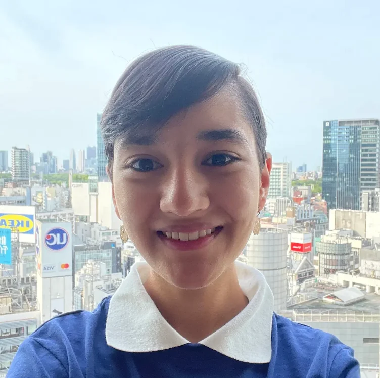 A young woman smiling in front of a city skyline with tall buildings and advertisements.