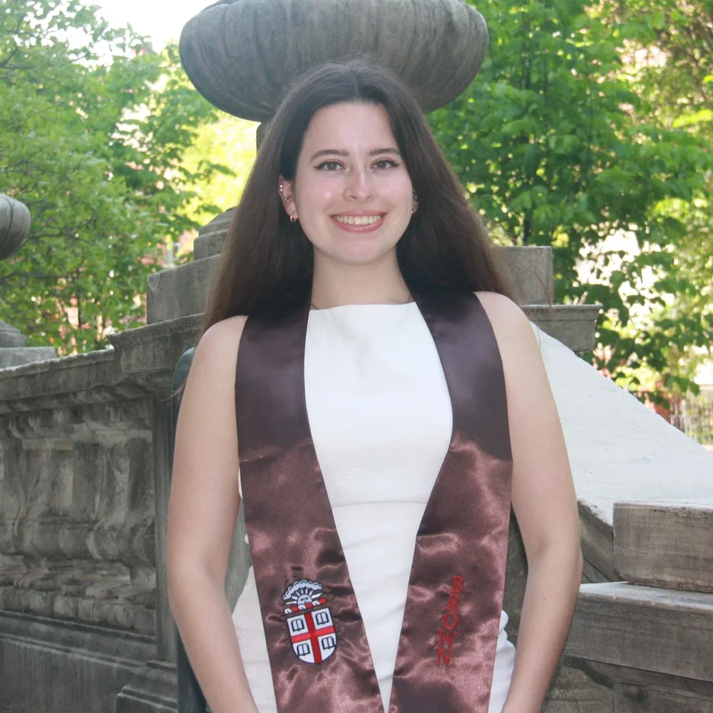 A man with glasses in a dark blue suit and white shirt, smiling outdoors with greenery in the background.