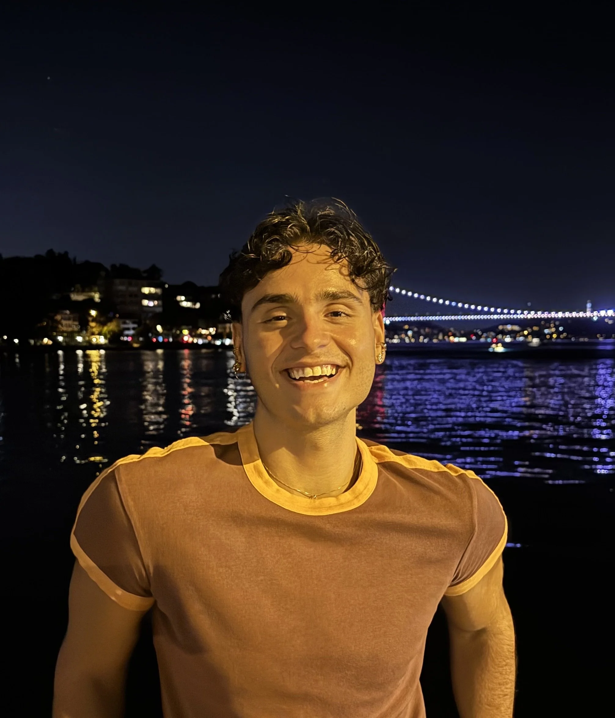 Smiling woman with short black hair, wearing a black polo shirt and jewelry, standing outdoors in front of a building with brick and a tree with autumn leaves.