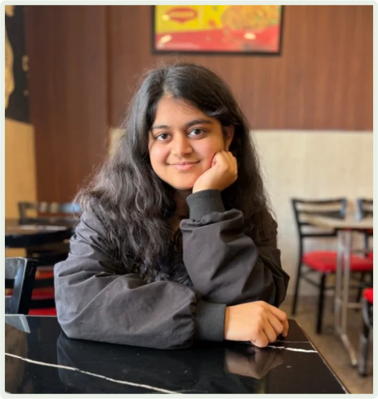 Young woman with long black hair sitting at a table in a restaurant, smiling and resting her chin on her hand.