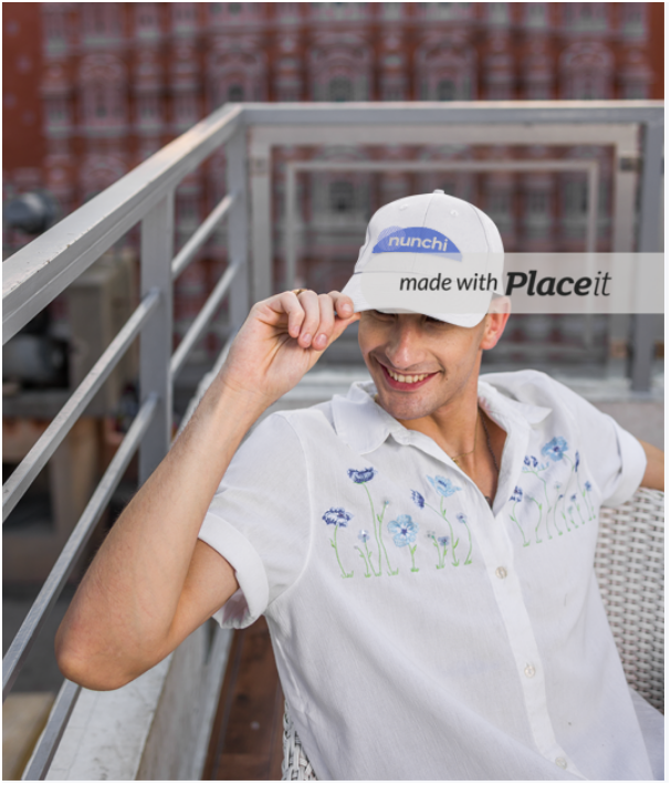 Man sitting on a rooftop balcony, wearing a white embroidered shirt and a white cap with the word 'nunichi' on it, smiling and adjusting his cap.