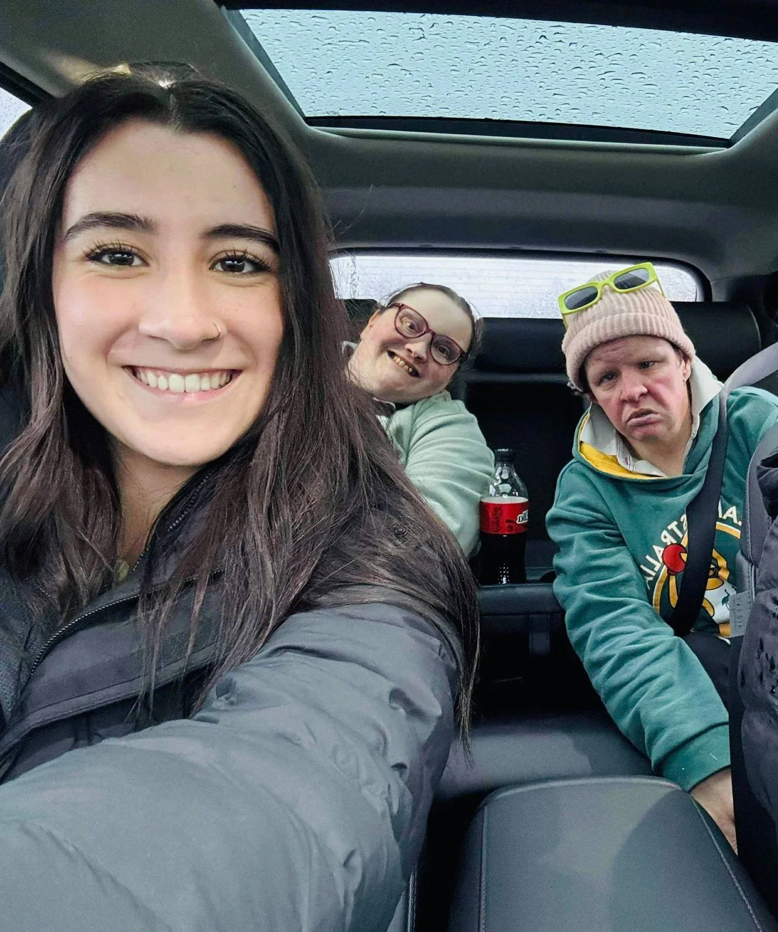 Three people sitting in a car, with one woman in the foreground smiling, and two others in the background with one wearing sunglasses on her hat and the other wearing glasses and smiling.