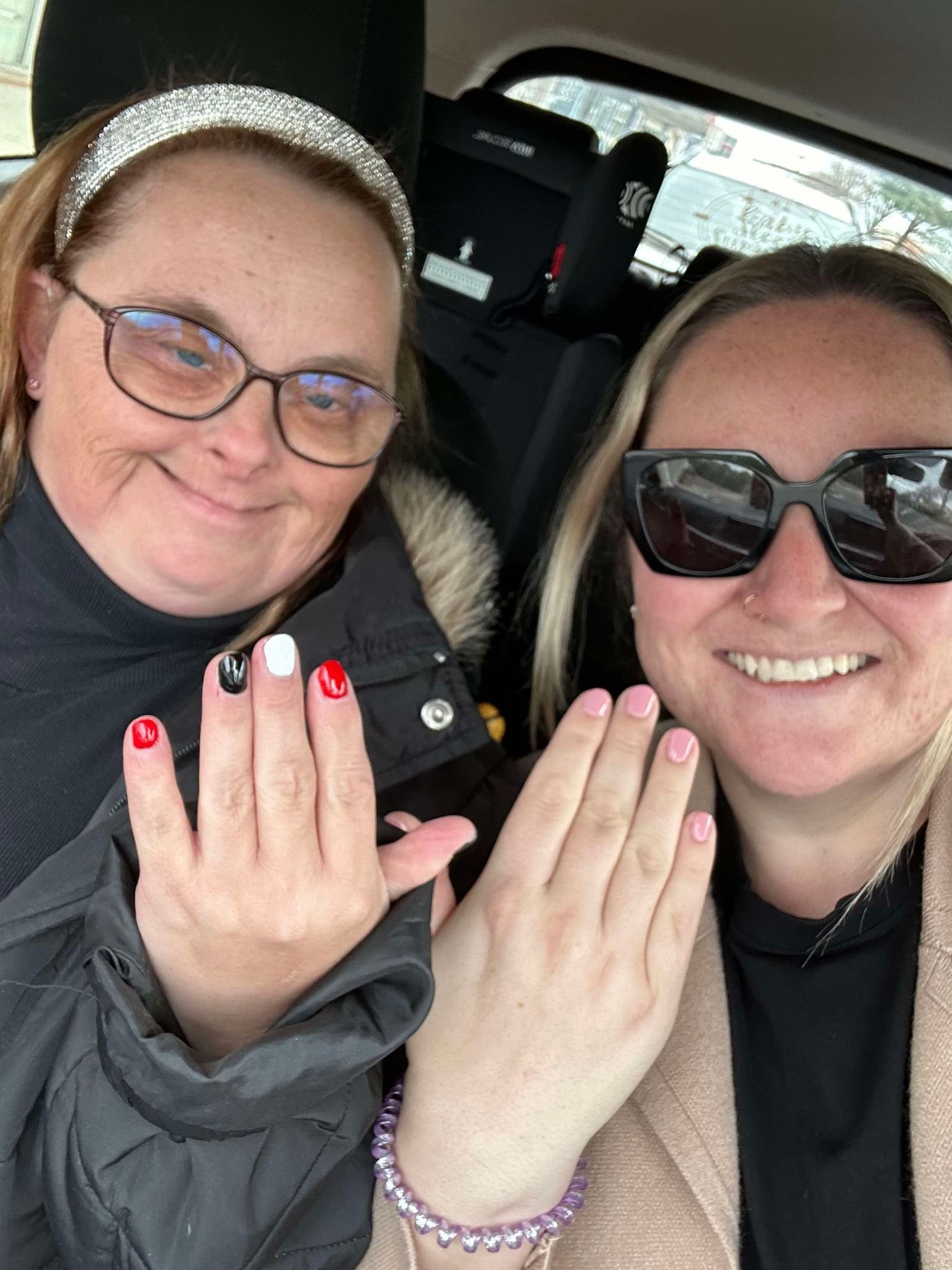 Two women showing their painted fingernails and wearing bracelets, taking a selfie inside a vehicle with a blurry view outside the window.