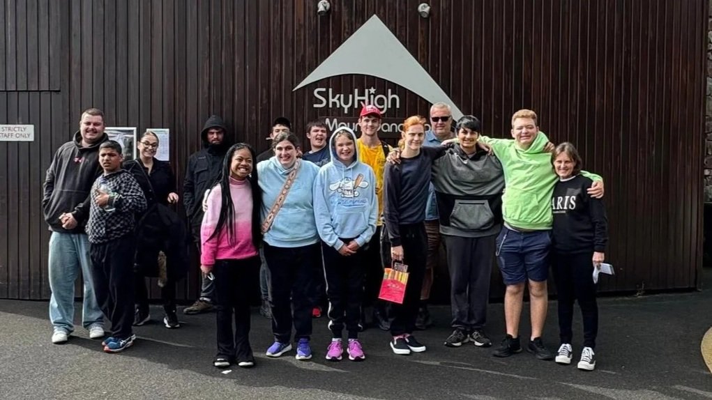 Group of people standing in front of a wooden wall with a sign that reads 'SkyHigh Moutain.'