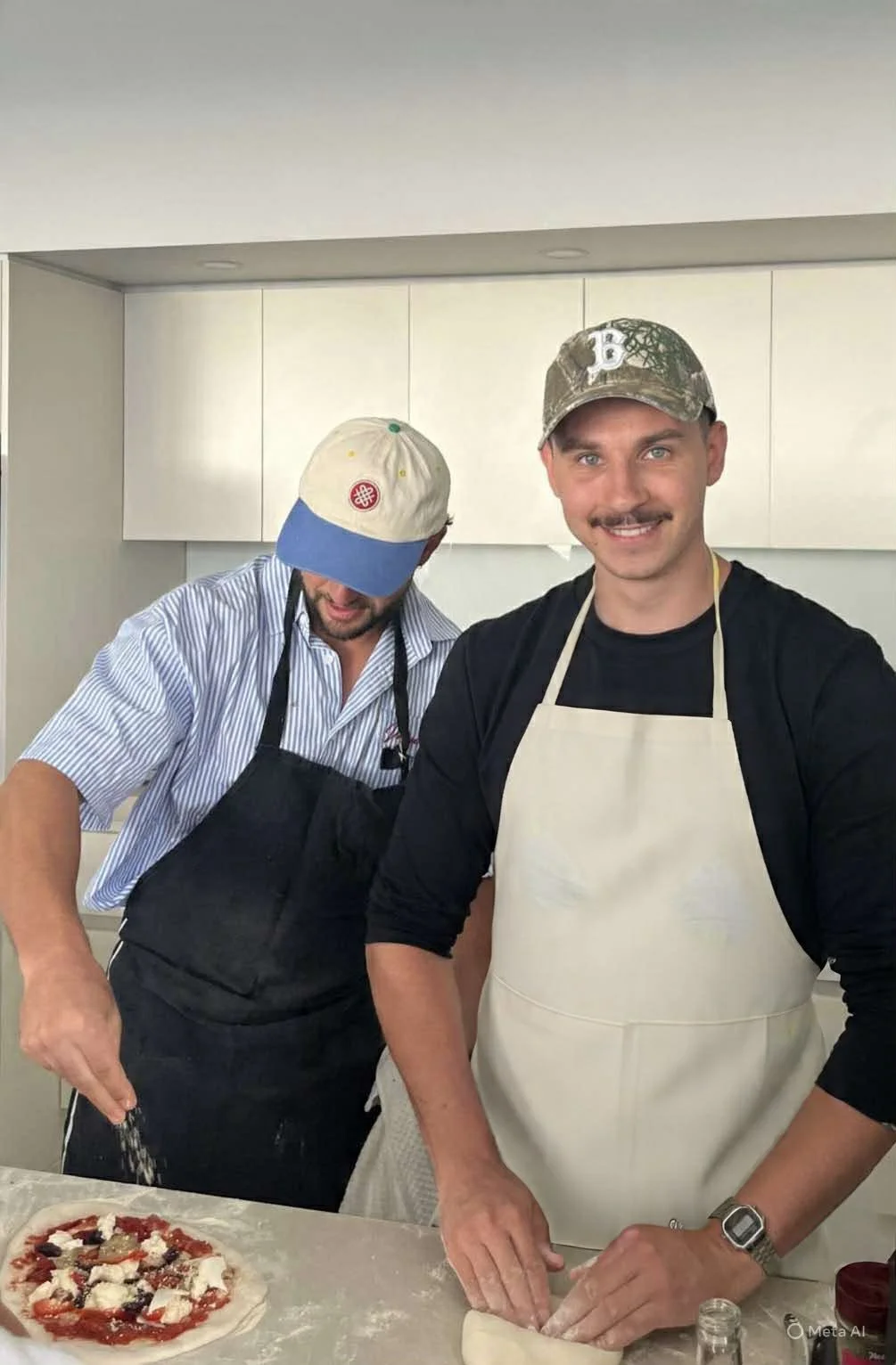 Two men preparing a pizza in a kitchen. The man on the right is smiling and wearing a camouflage cap and an apron. The man on the left is looking down, wearing a baseball cap and an apron. There is a pizza with toppings on the counter in front of them.