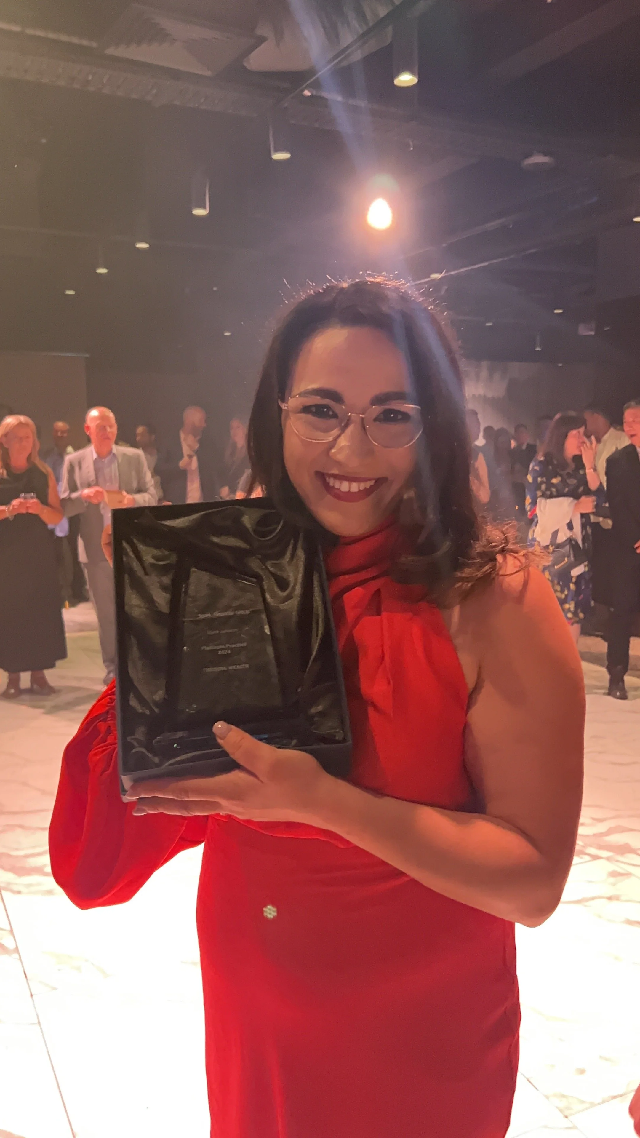 A woman with glasses and long dark hair in a red dress is smiling and holding an award plaque in a social event or party setting with people in the background.