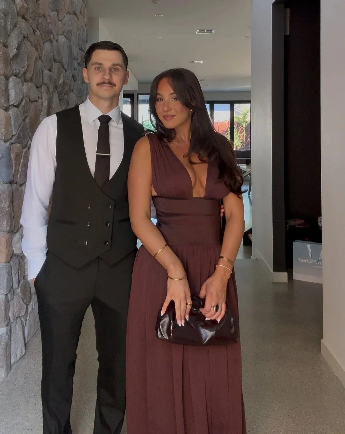 A man and woman standing indoors, dressed in formal attire, with a stone wall on the left and large windows in the background showing a patio and outdoor plants.