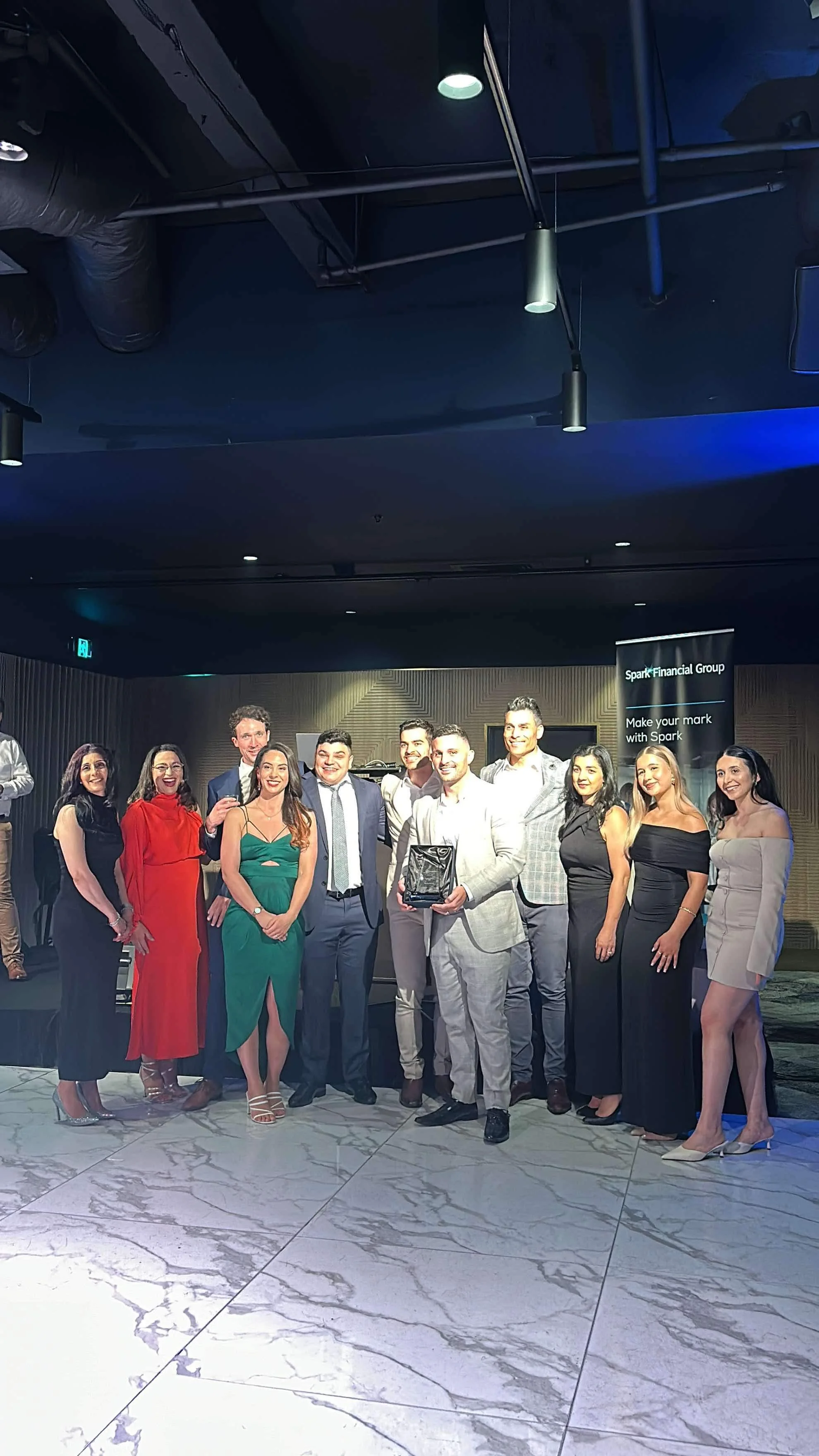 Group of people at a formal celebration or award ceremony, posing on stage with one person holding a plaque or award, dressed in suits and evening wear, in a venue with marble flooring and a banner in the background.