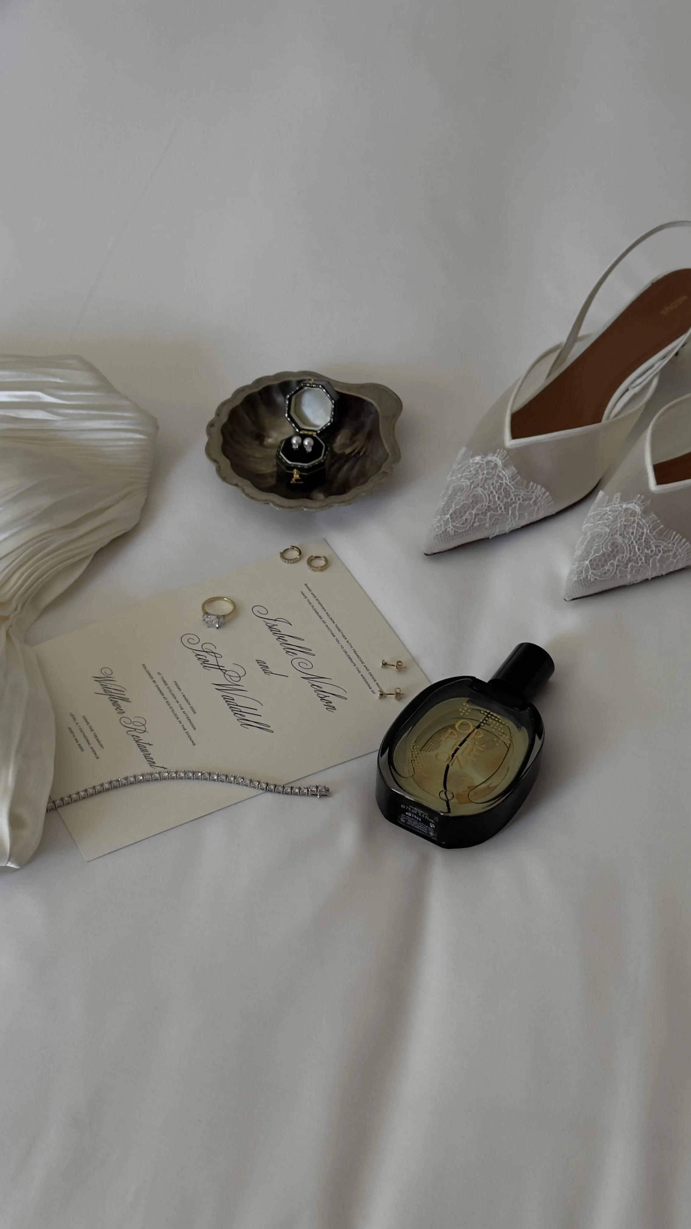 Wedding accessories including rings, earrings, bracelet, perfume, white lace shoes, and invitation on a white surface.