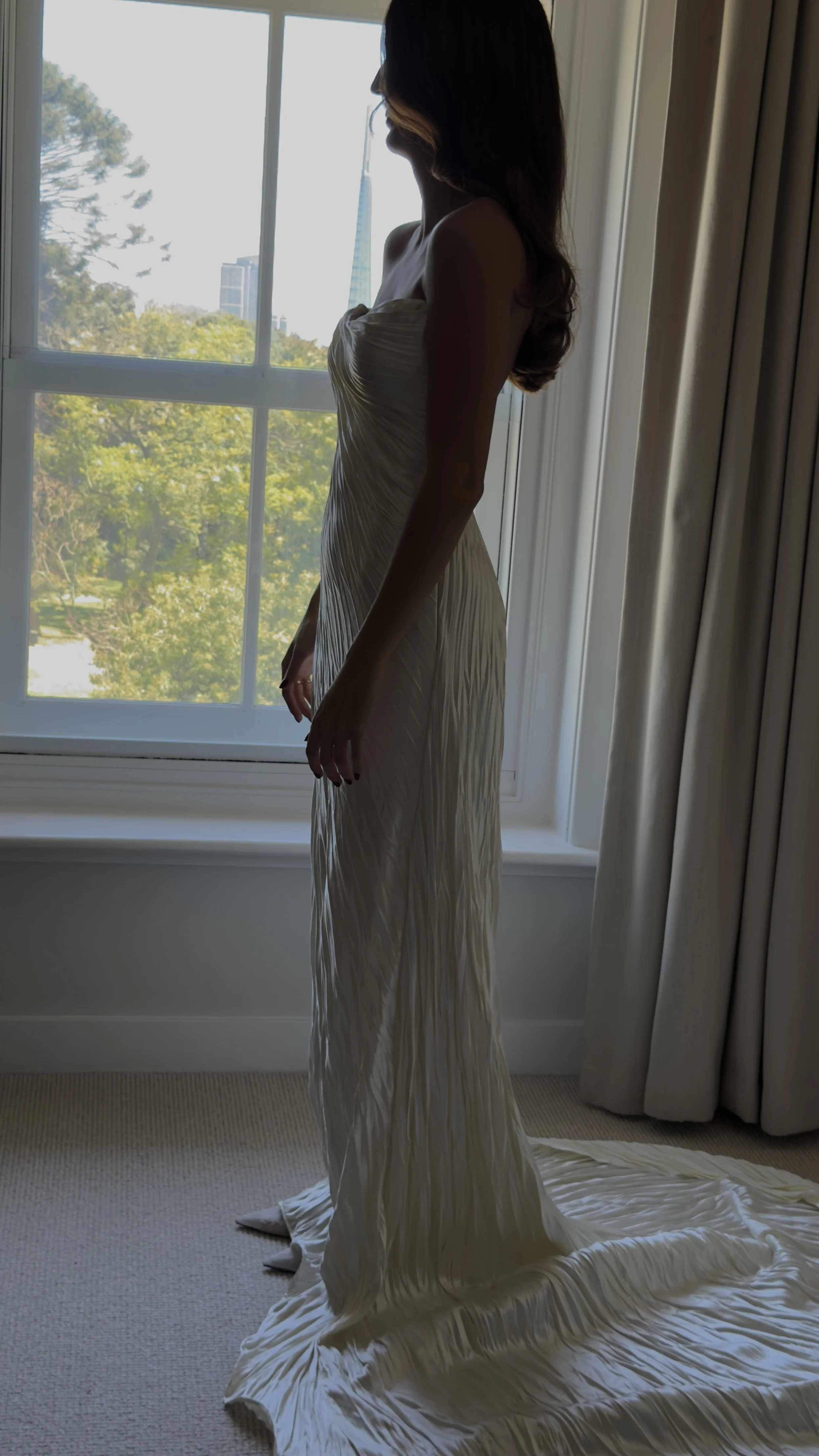 A woman standing inside near a window with trees outside, wearing a long, gathered white dress, with the train spread on the carpeted floor.
