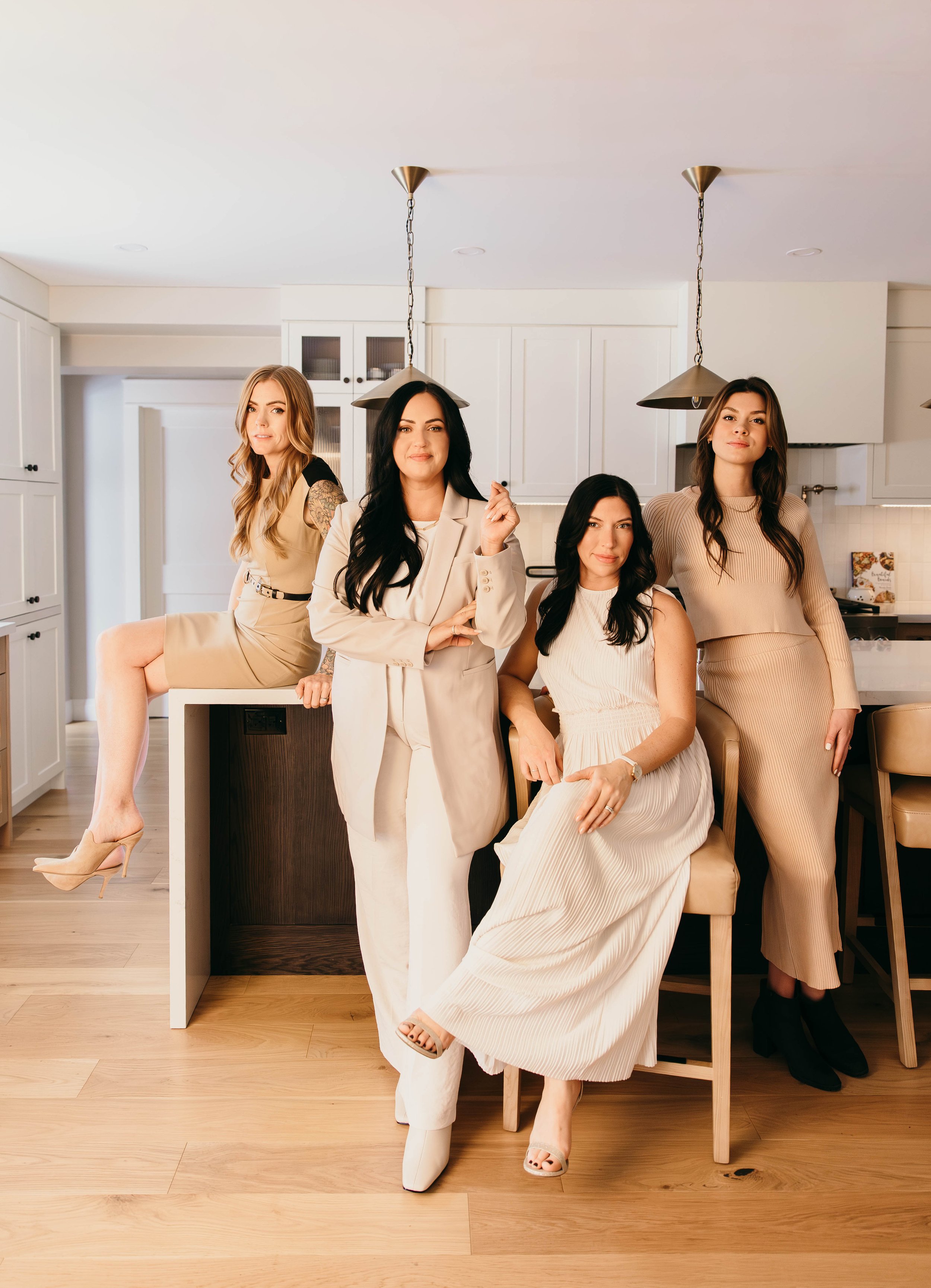 Four women in neutral-colored outfits posing in a modern kitchen, with one sitting on a counter and the others standing around a barstool.