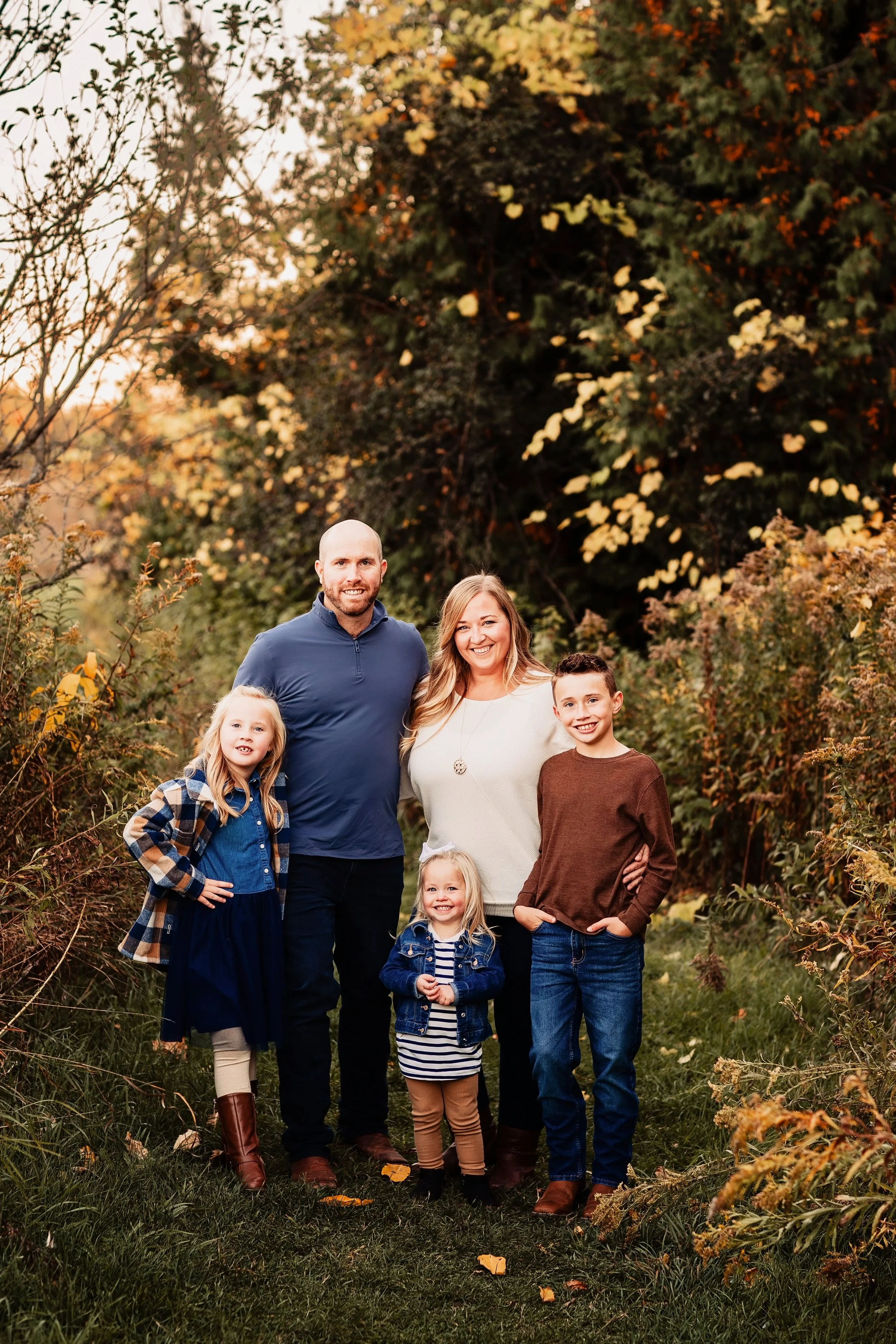 A family of six posing outdoors on a fall day with colorful autumn trees in the background.