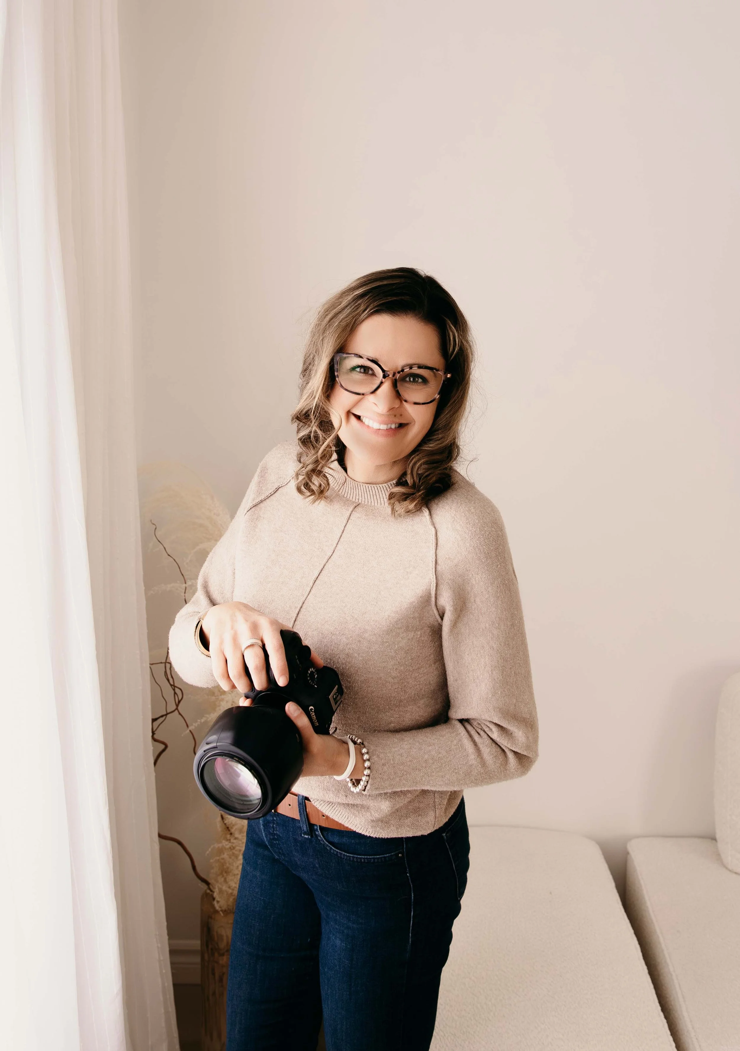 A woman with wavy brown hair, wearing glasses and a beige sweater, smiling and holding a professional camera inside a room with neutral-colored decor. Morgan Bress Photography