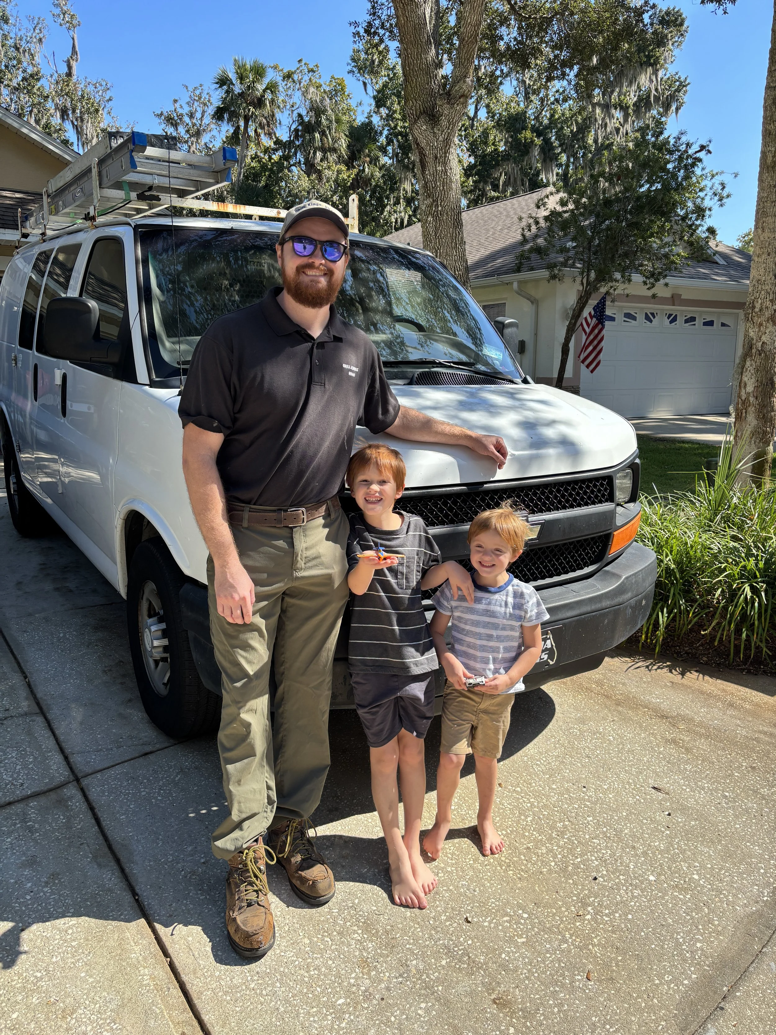 A man with a beard, sunglasses, and a cap standing next to two young boys in front of a white van on a sunny day. The boys are holding toy cars, and they are all smiling.