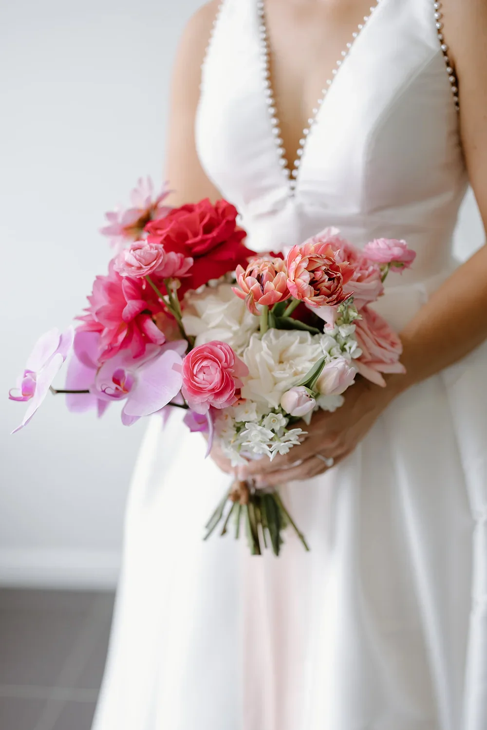 Bridal bouquet with pink roses, tulips, orchids, ranunculus with white modern bridal dress in the background.