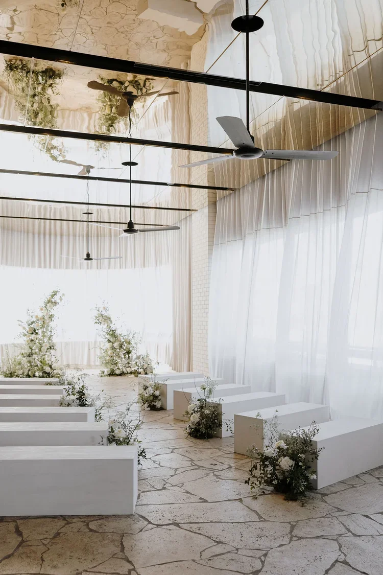 Modern white wedding ceremony setup with lush garden-style floral pillars and aisle arrangements at The Calile in Brisbane.