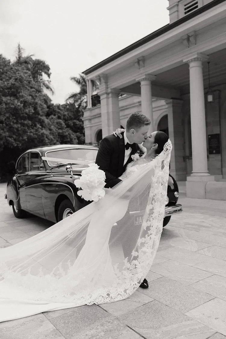 Bride and groom sharing a romantic dip and kiss beside a vintage black wedding car, with the bride’s long lace veil flowing across the pavement.