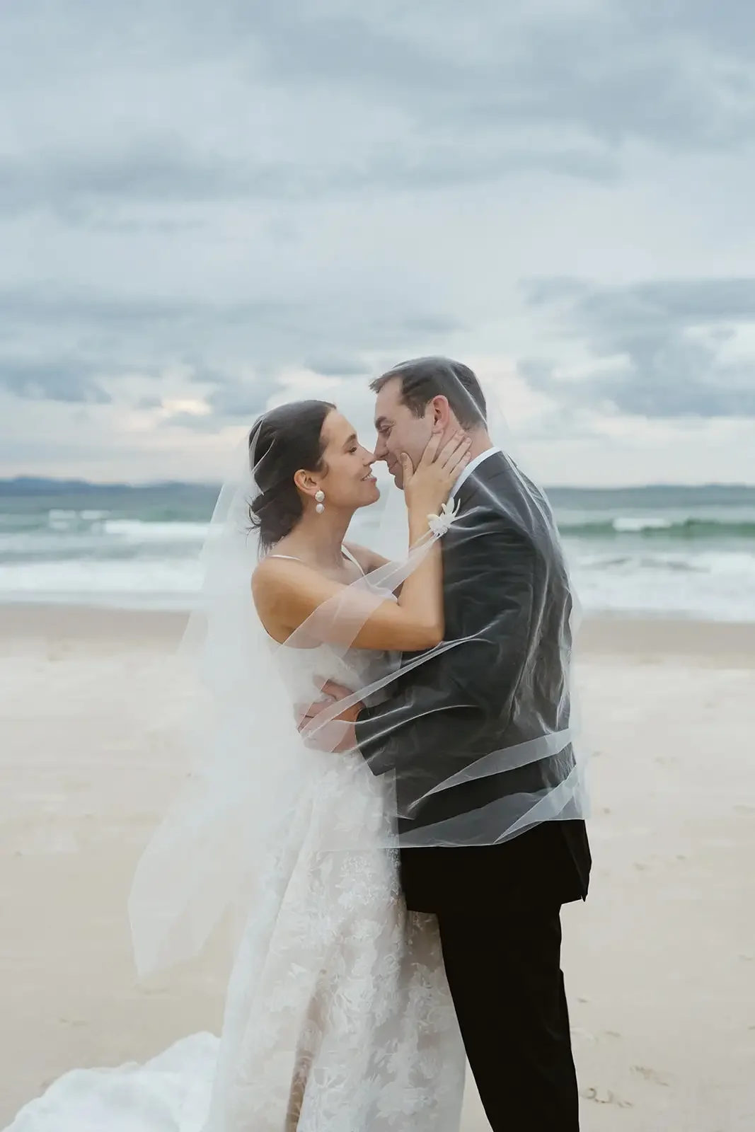 Bride and groom embracing on Byron Bay beach with veil blowing in the wind.