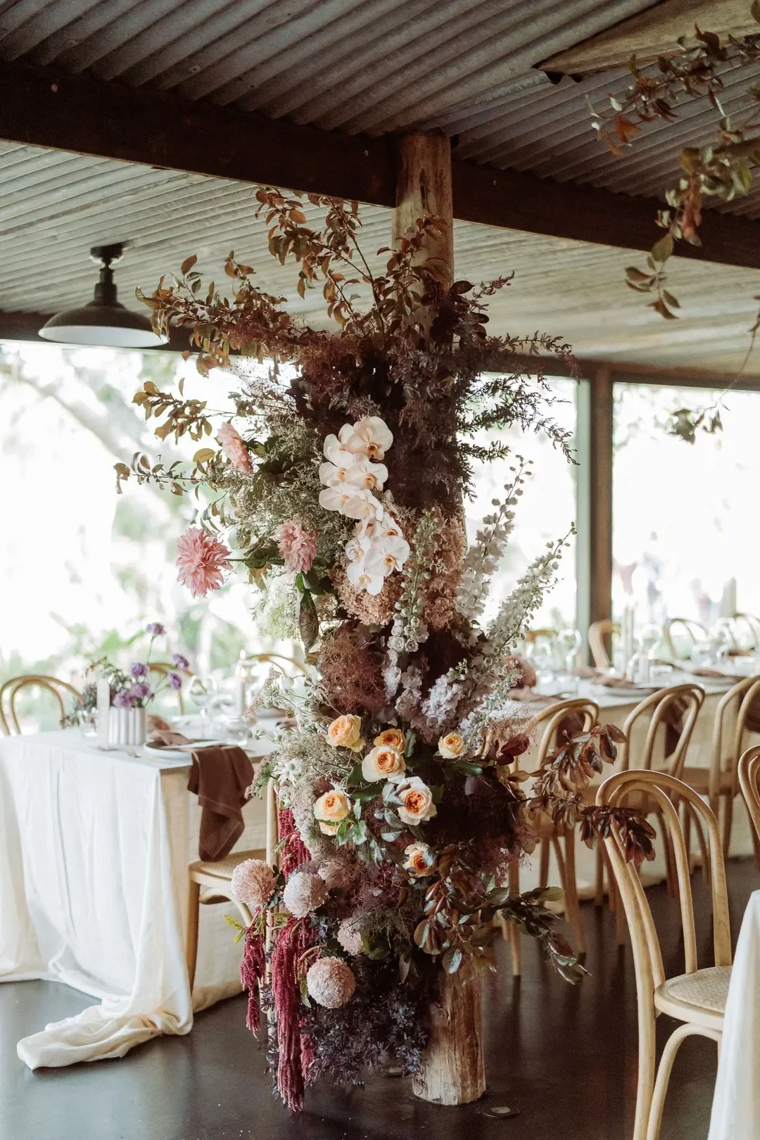 Large floral installation on a timber post featuring roses, orchids, smoke bush, and cascading textural blooms.