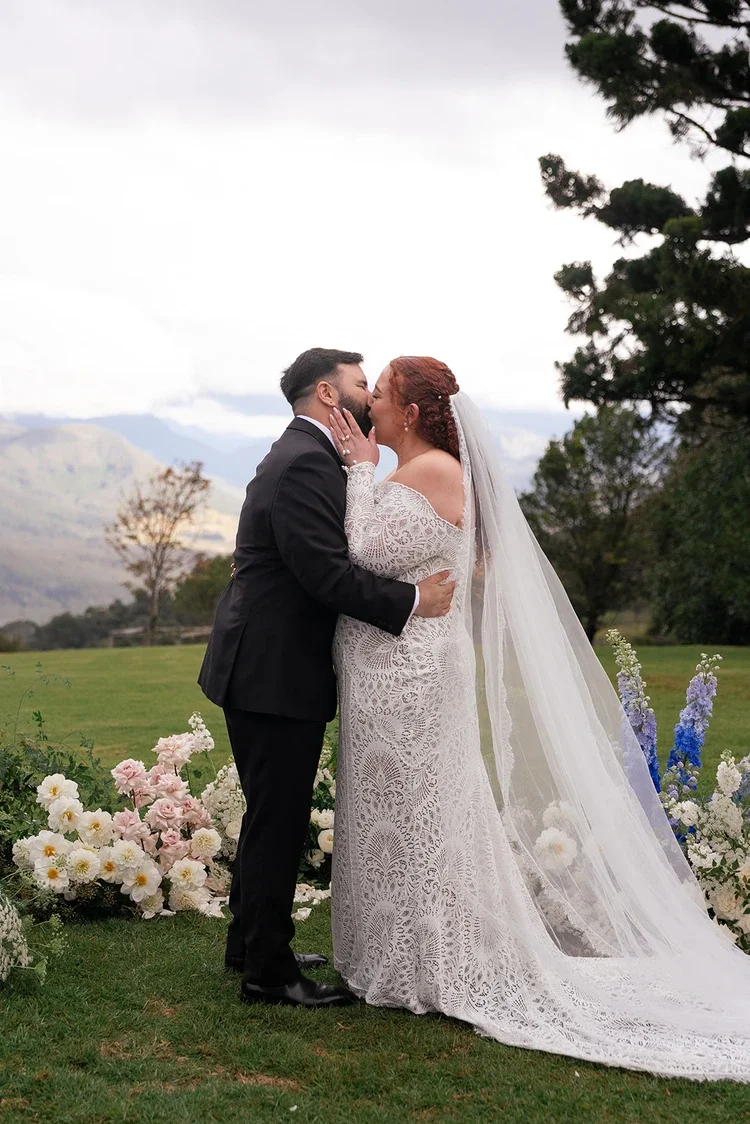 Bride and groom sharing their first kiss surrounded by lush white, pink, and blue floral arrangements with mountains in the background.”