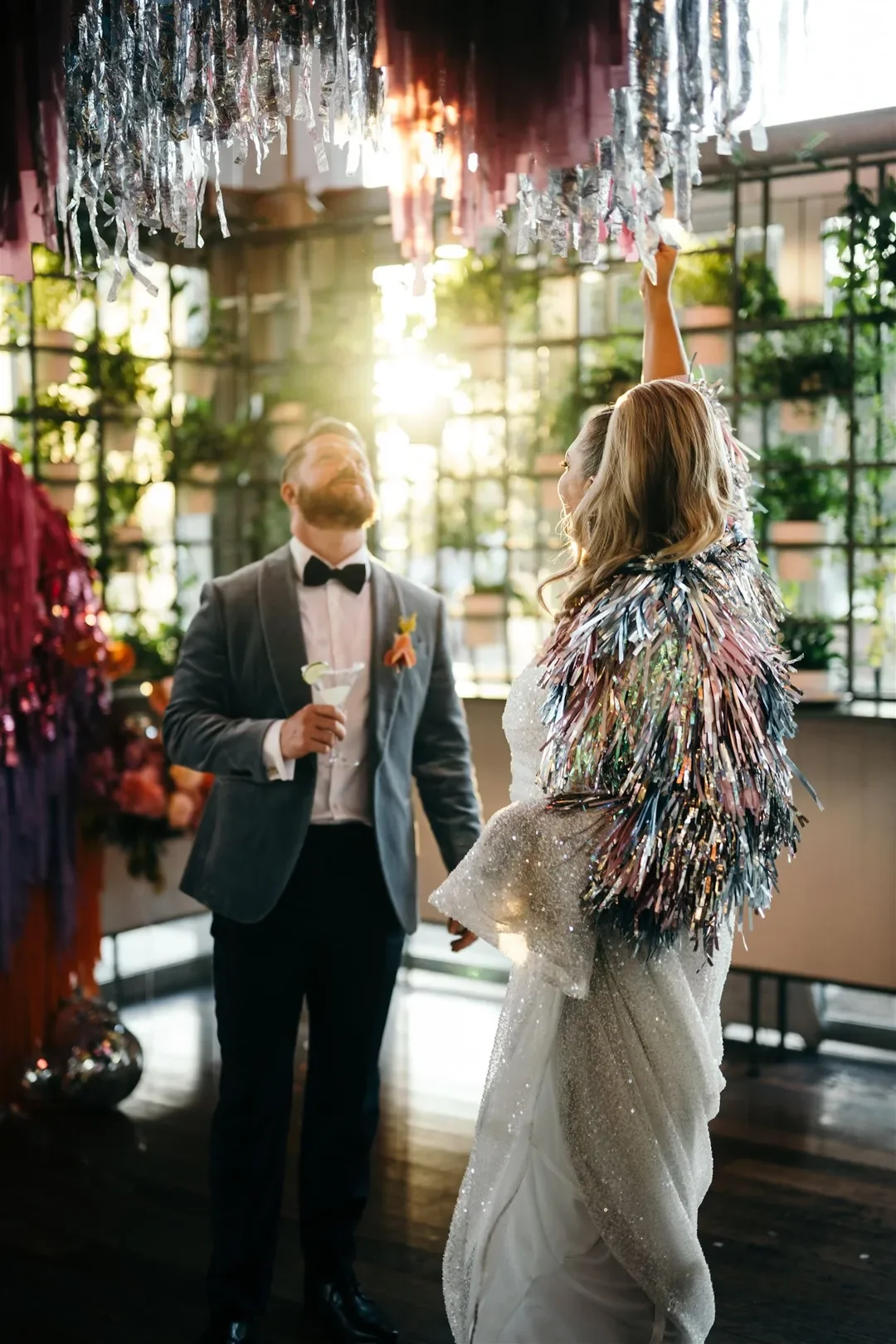 Sarah and Marshall standing beneath hanging streamers as sunlight shines behind them during their fun and vibrant wedding reception.