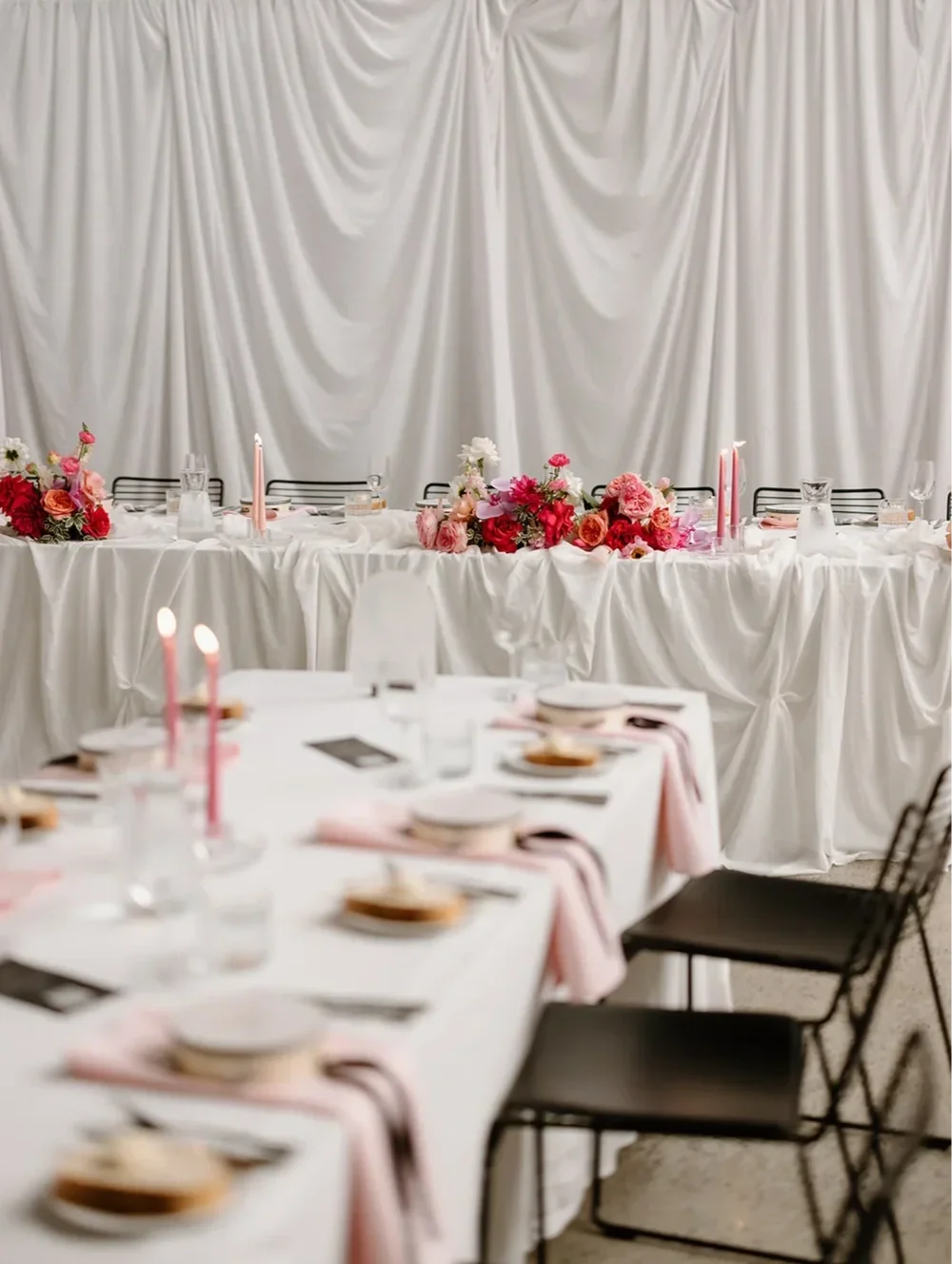 Vibrant table arrangement with bold pink ranunculus, hot pink dahlias, and white florals against white draping.