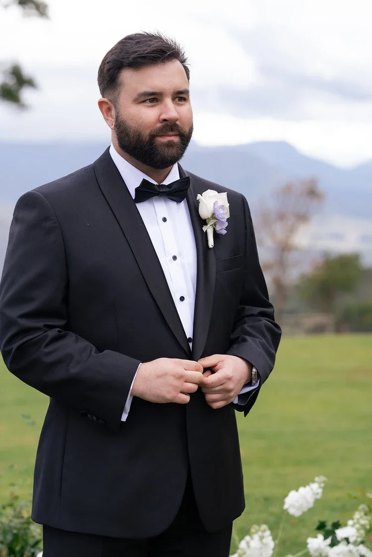 Groom standing outdoors in a classic black tuxedo with a white and lilac boutonnière against a scenic mountain backdrop.