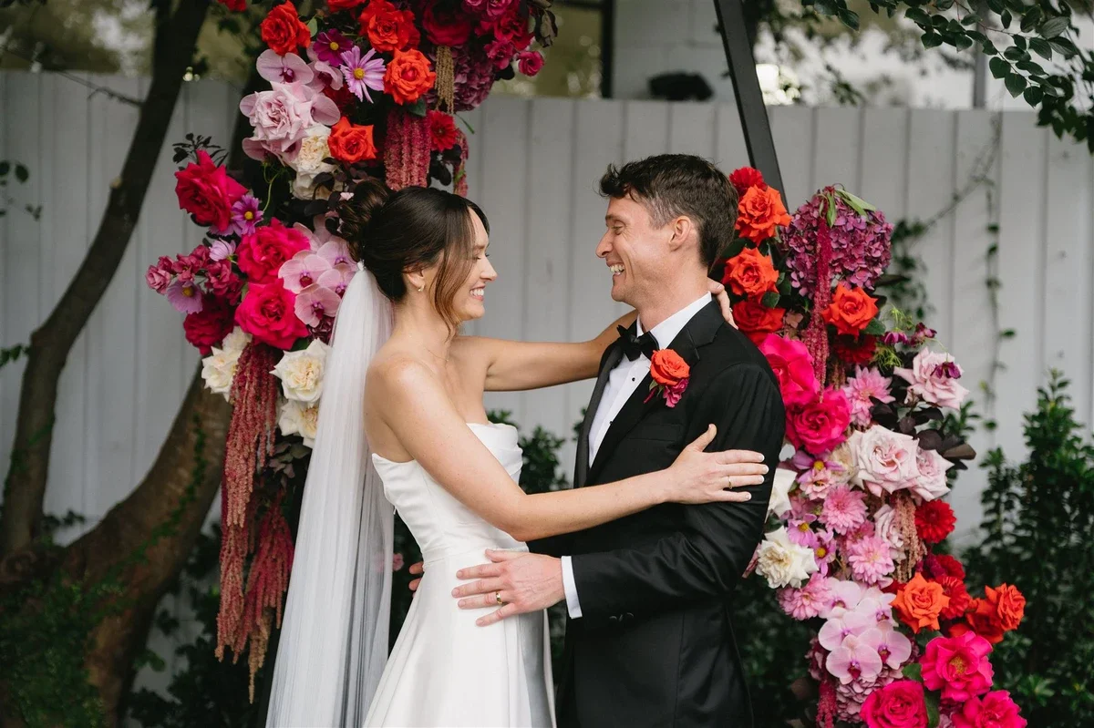 Smiling couple framed by vivid pink, peach and  red roses and dahlias on the ceremony arbour.