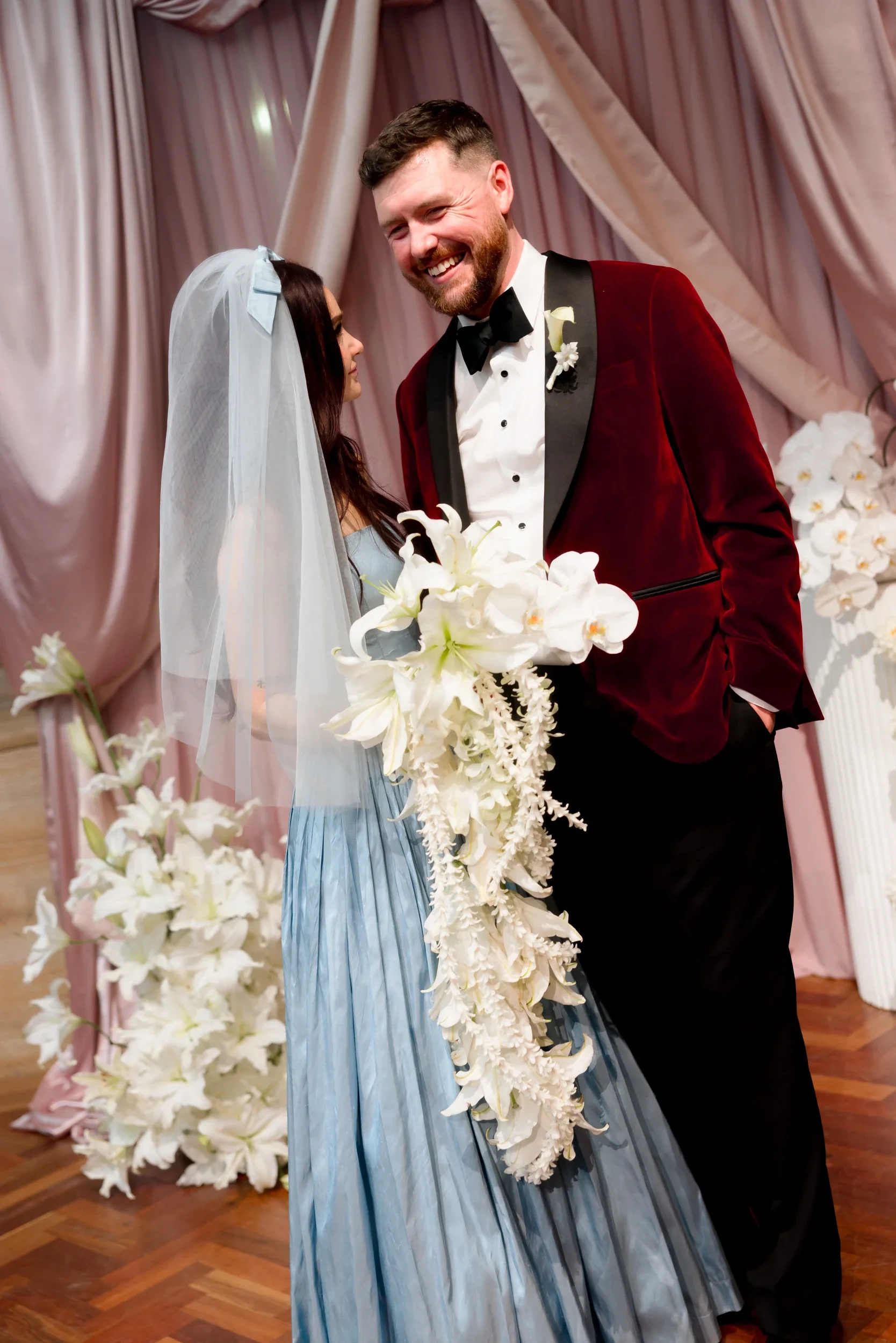 Bride holding a long trailing white bouquet beside the groom in burgundy velvet