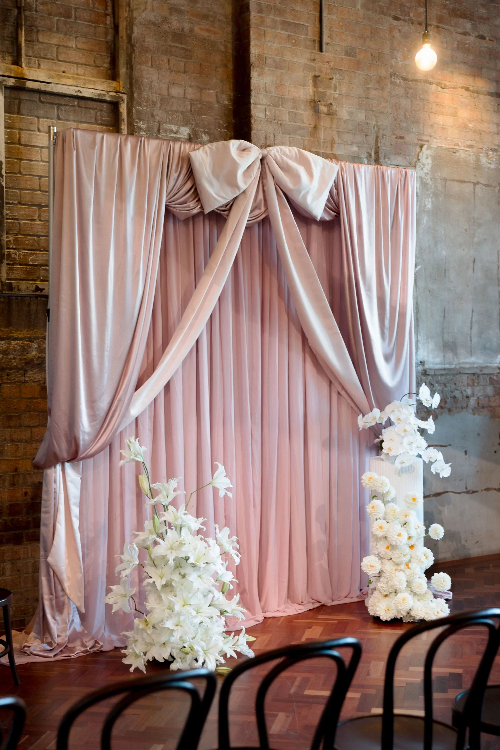 Blush draped ceremony backdrop with an oversized bow, framed by white lily and orchid florals at The Warehouse Brisbane
