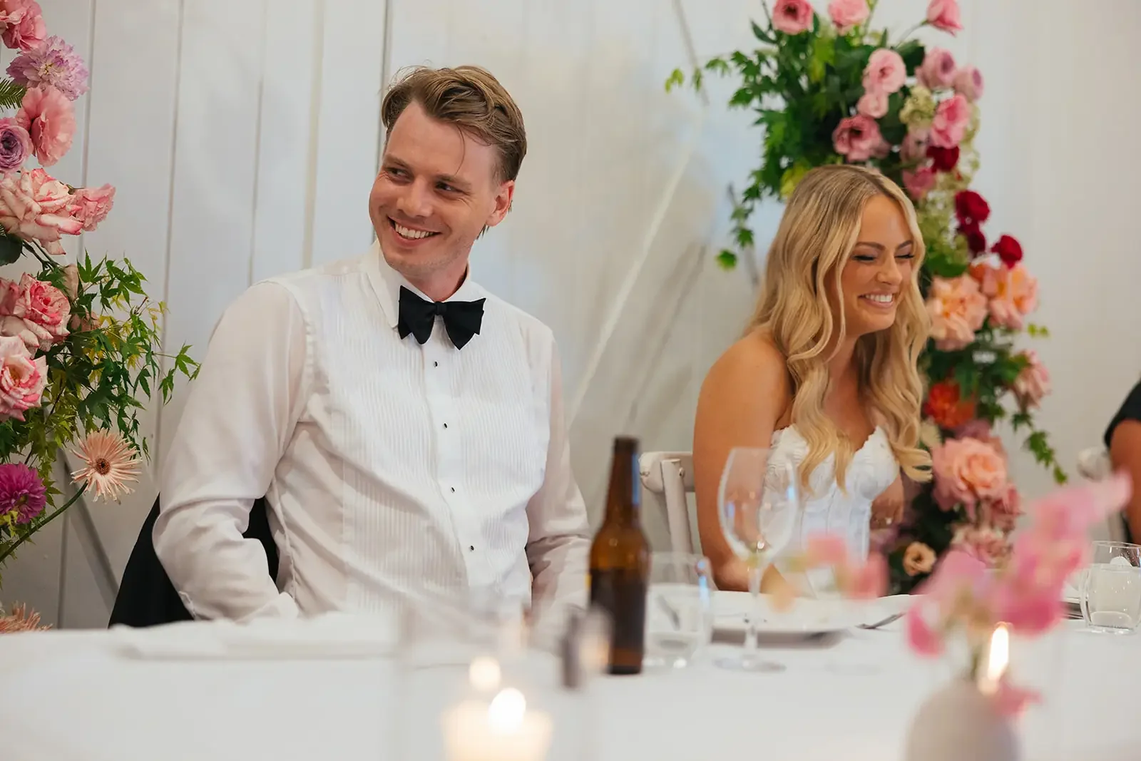 Wedding couple seated at their reception table, smiling with vibrant pink and peach floral arrangements surrounding them.